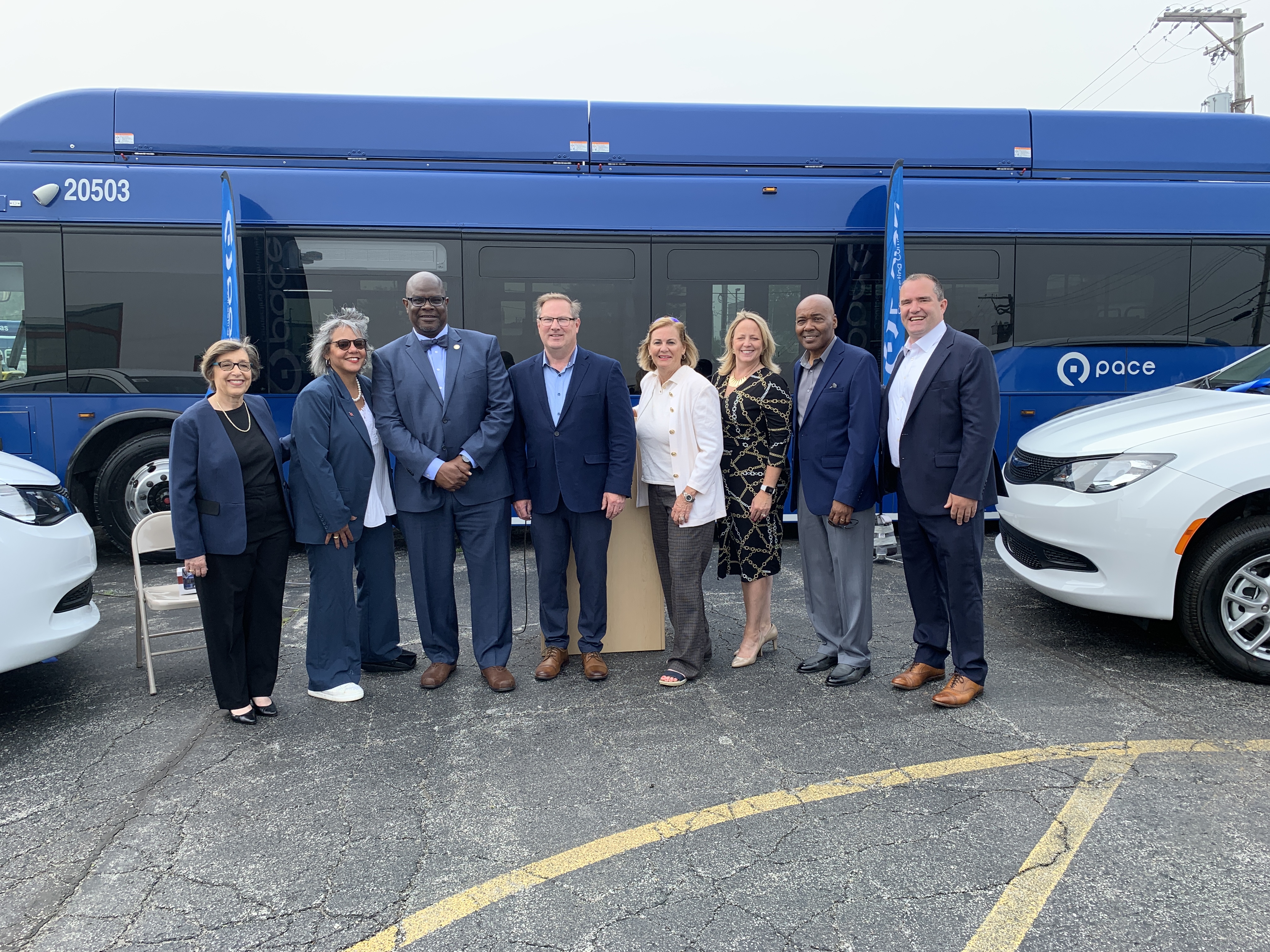 An image of officials at Pace's Harvey Transportation Center. The image shows Pace Executive Director Melinda Metzger; Congresswoman Robin Kelly; Harvey Mayor Christopher J Clark; Pace Chairman Rick Kwasneski; RTA Board Member Elizabeth Doody Gorman; Cook County Department of Transportation and Highways (DoTH) Superintendent Jennifer “Sis” Killen; Pace Director Terry R. Wells; and Pace Director David Guerin in front of a Pace bus and next to Pace Connect vans.