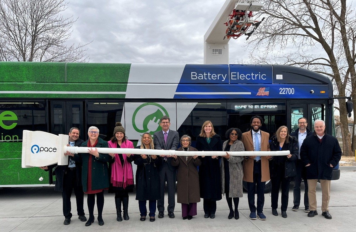 Group of legislators and Pace officials standing in front of a bus while holding an oversized electric plug.
