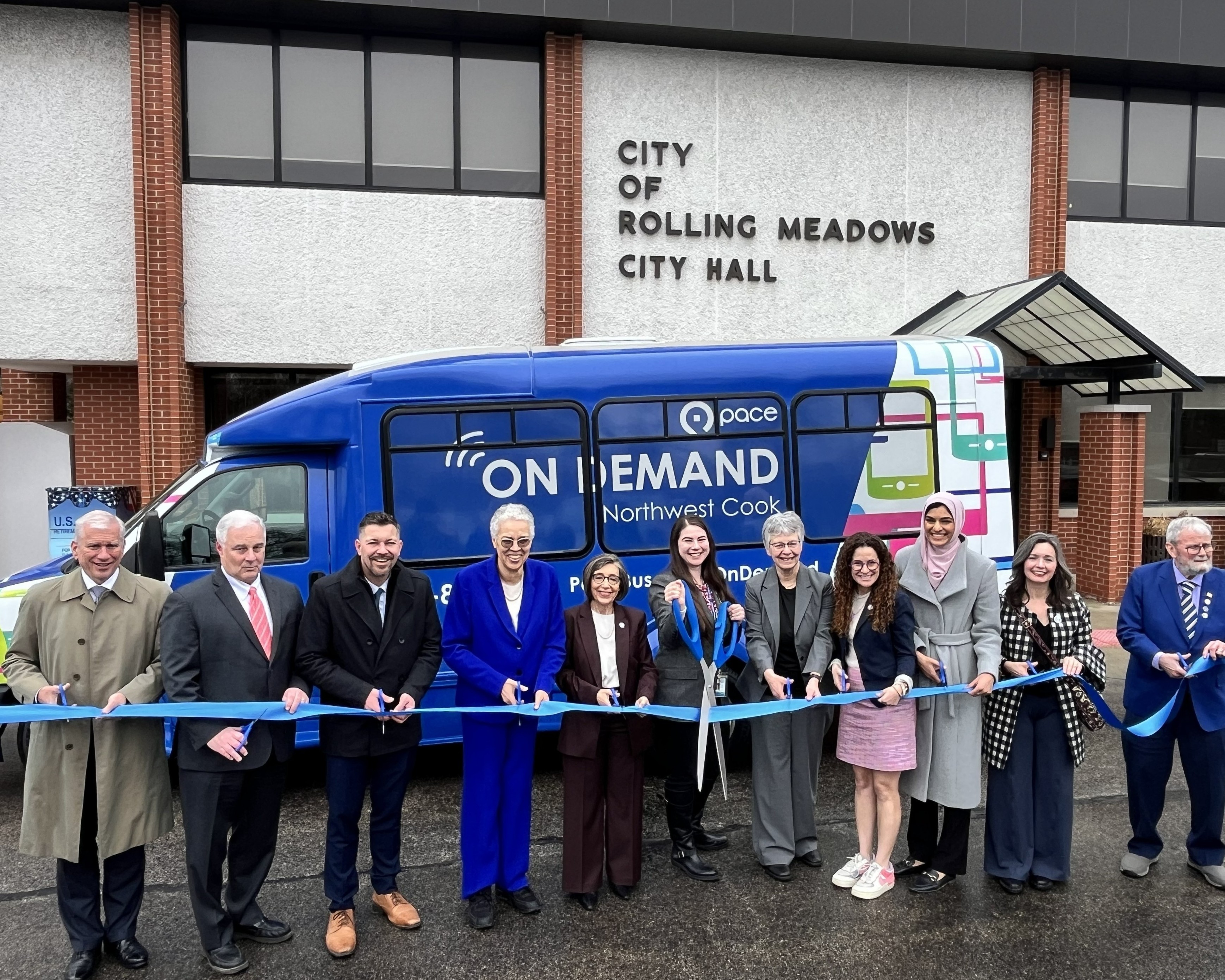 Photo of elected officials and Pace representatives standing in front of an On Demand bus and cutting a blue ribbon with scissors