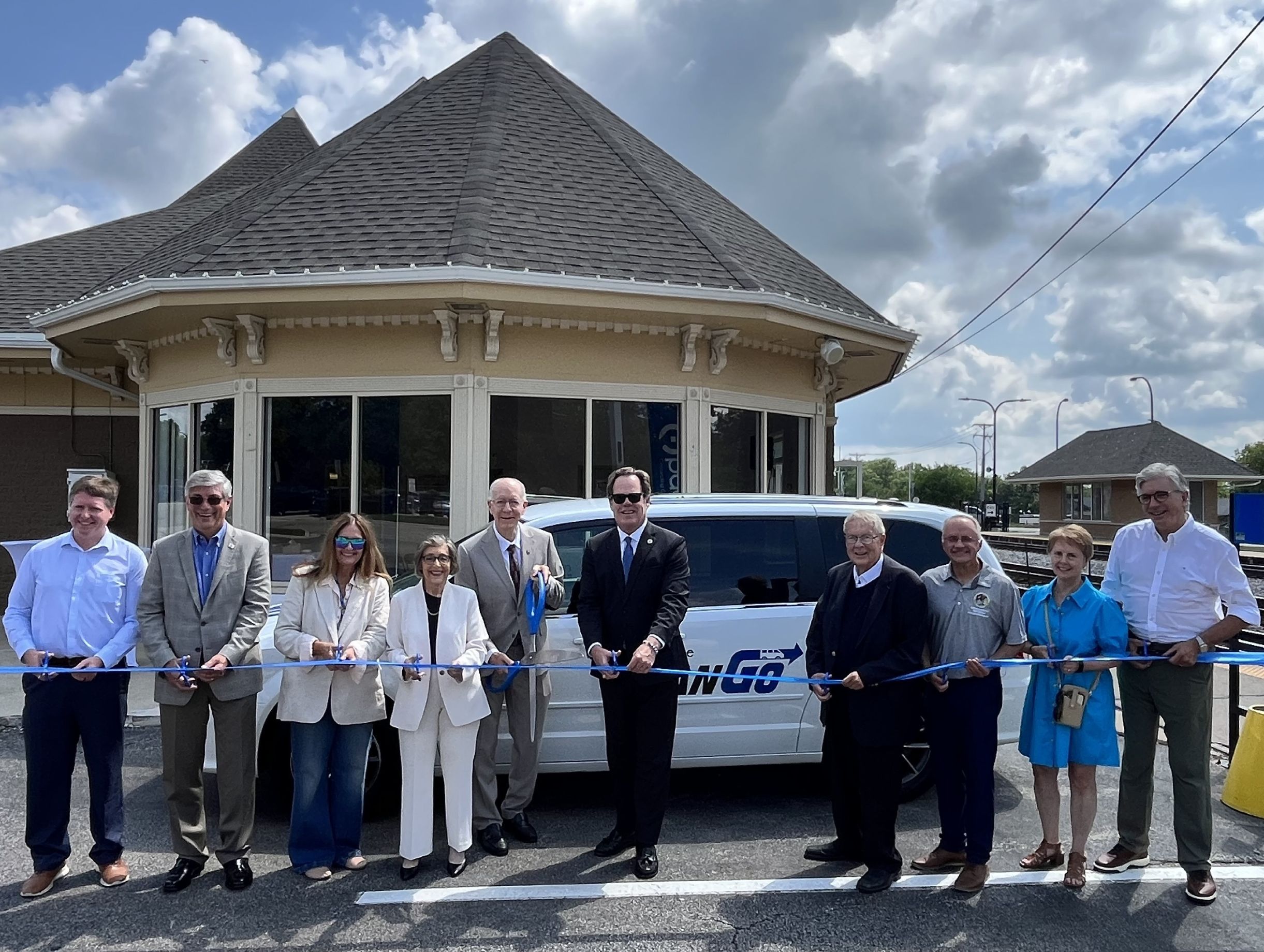 Photo of officials in Geneva and Pace staff members cutting a blue ribbon in front of a VanGo van and standing outside at the Geneva Metra Station